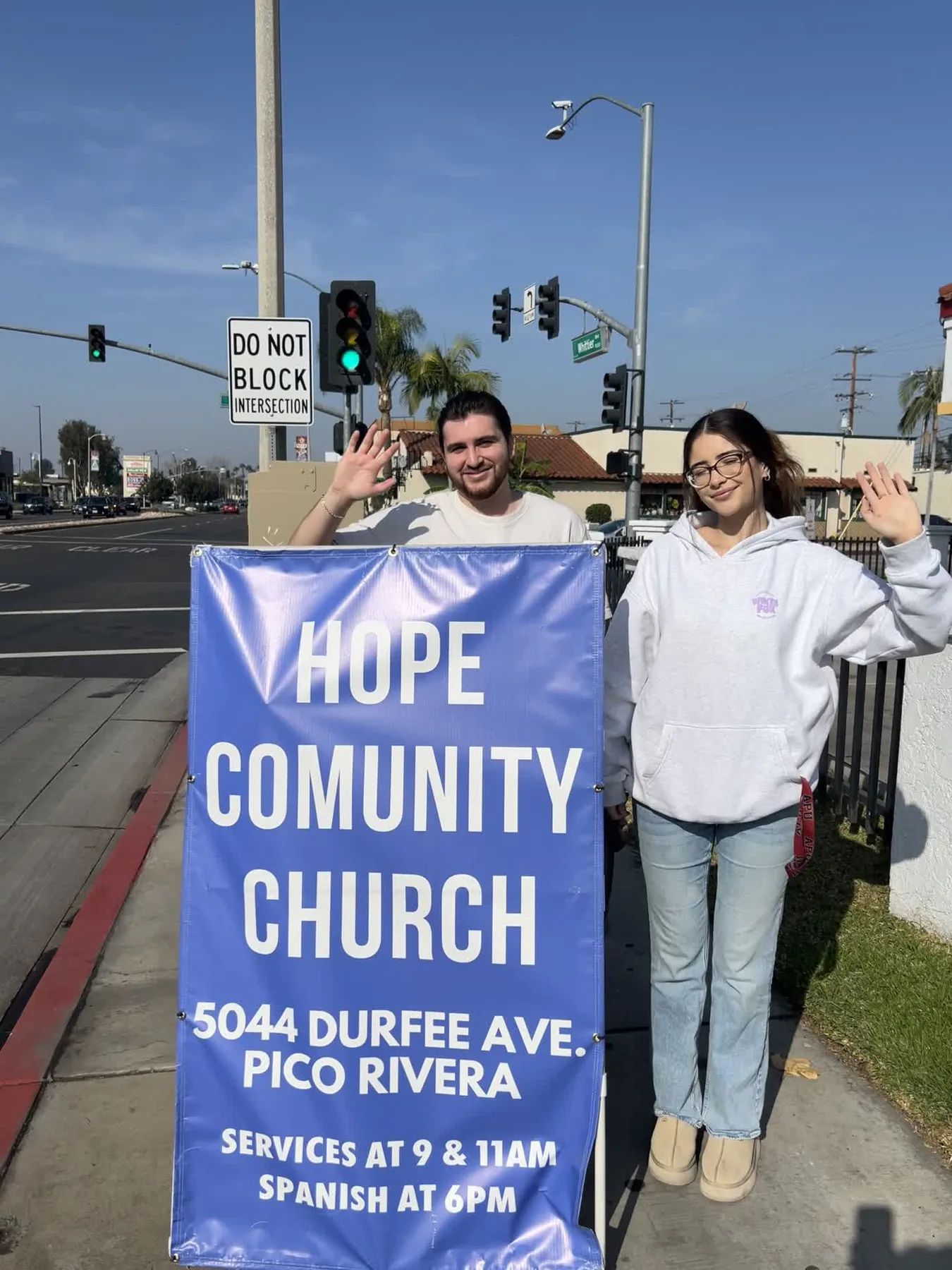 Two Hope Community Church members smiling and waving beside a blue Sign Ministry sign